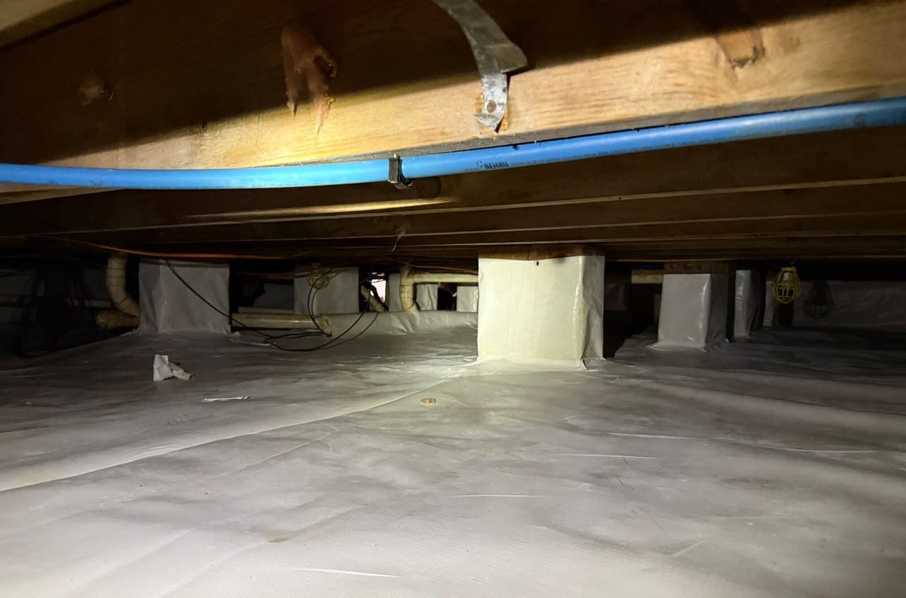 Lined crawl space floor with several wrapped piers under wooden joists and a blue water line.
