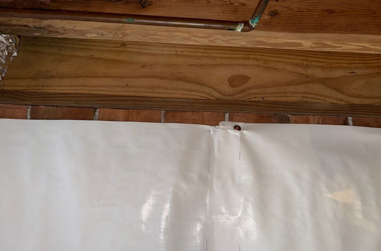 White crawl space wall liner fastened below the sill with a taped seam and a copper pipe above.
