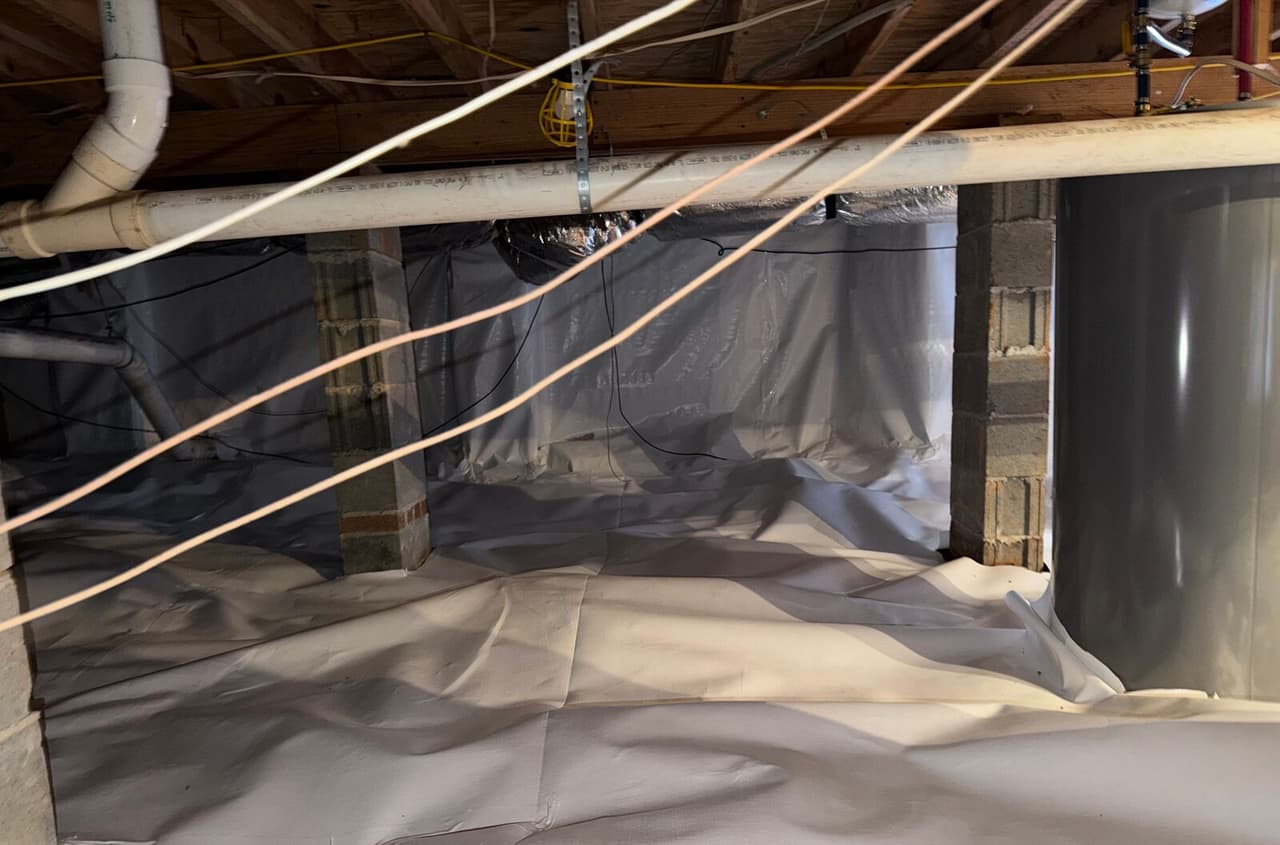 Crawl space with white liner covering the floor, walls and block piers under plumbing pipes.