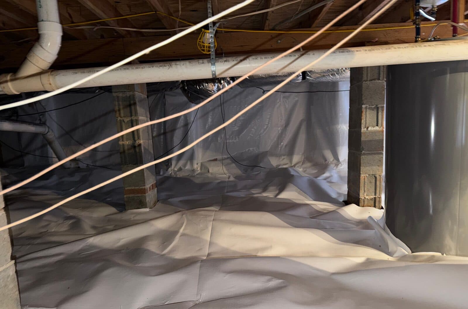 Crawl space with white liner covering the floor, walls and block piers under plumbing pipes.