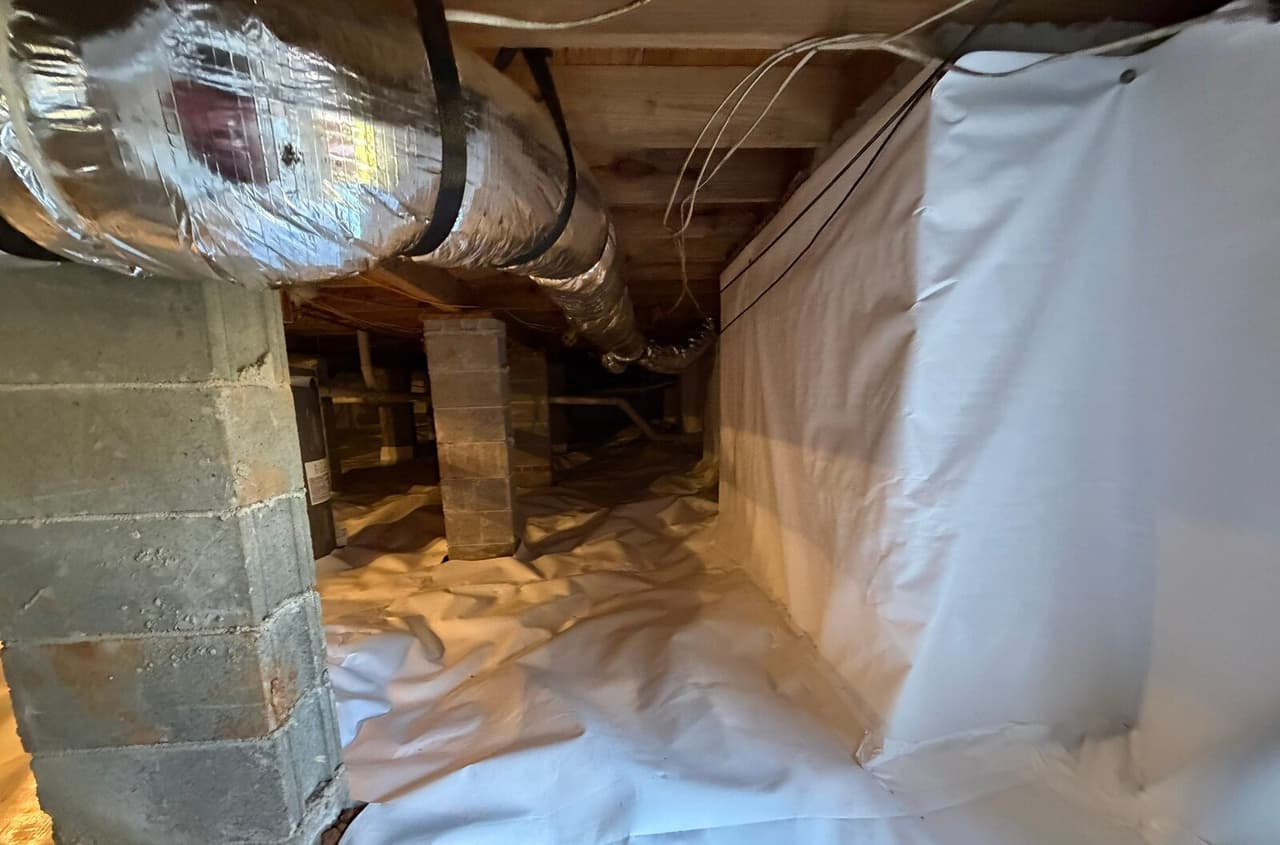 Narrow crawl space with white liner on the wall and floor beside a block pier and duct.