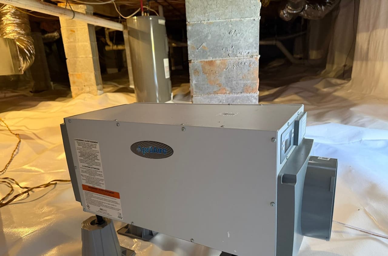 Dehumidifier on stands over white crawl space liner with piers and a water heater behind.