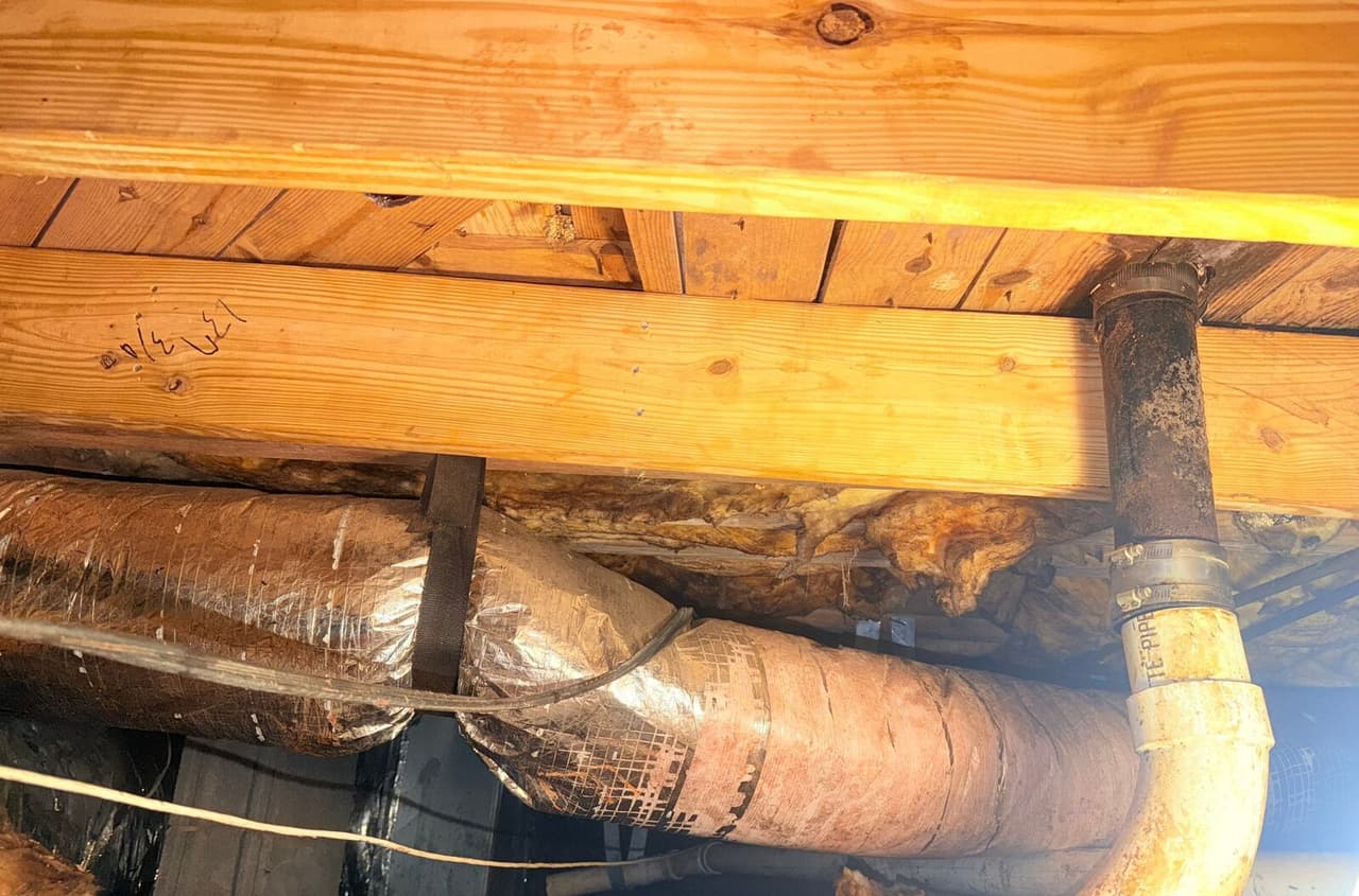 New lumber beam nailed under floor joists above insulated ductwork and a drain pipe.