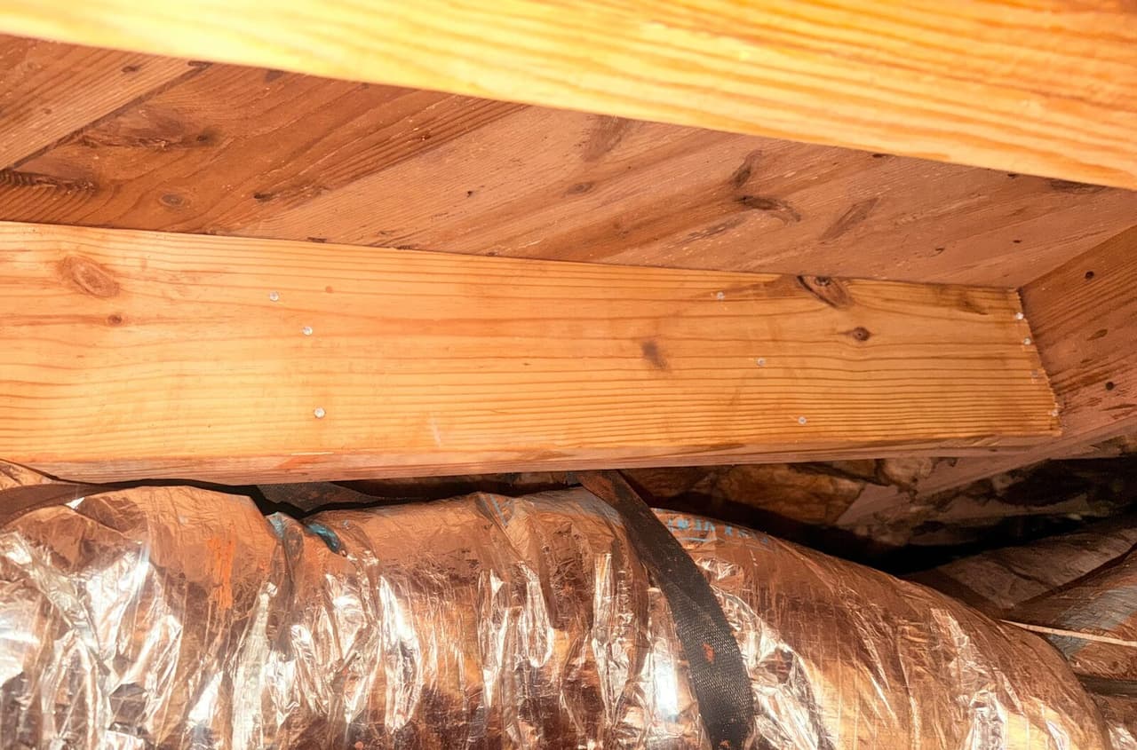 New wood board nailed beneath a subfloor above foil wrapped ductwork in a crawl space.