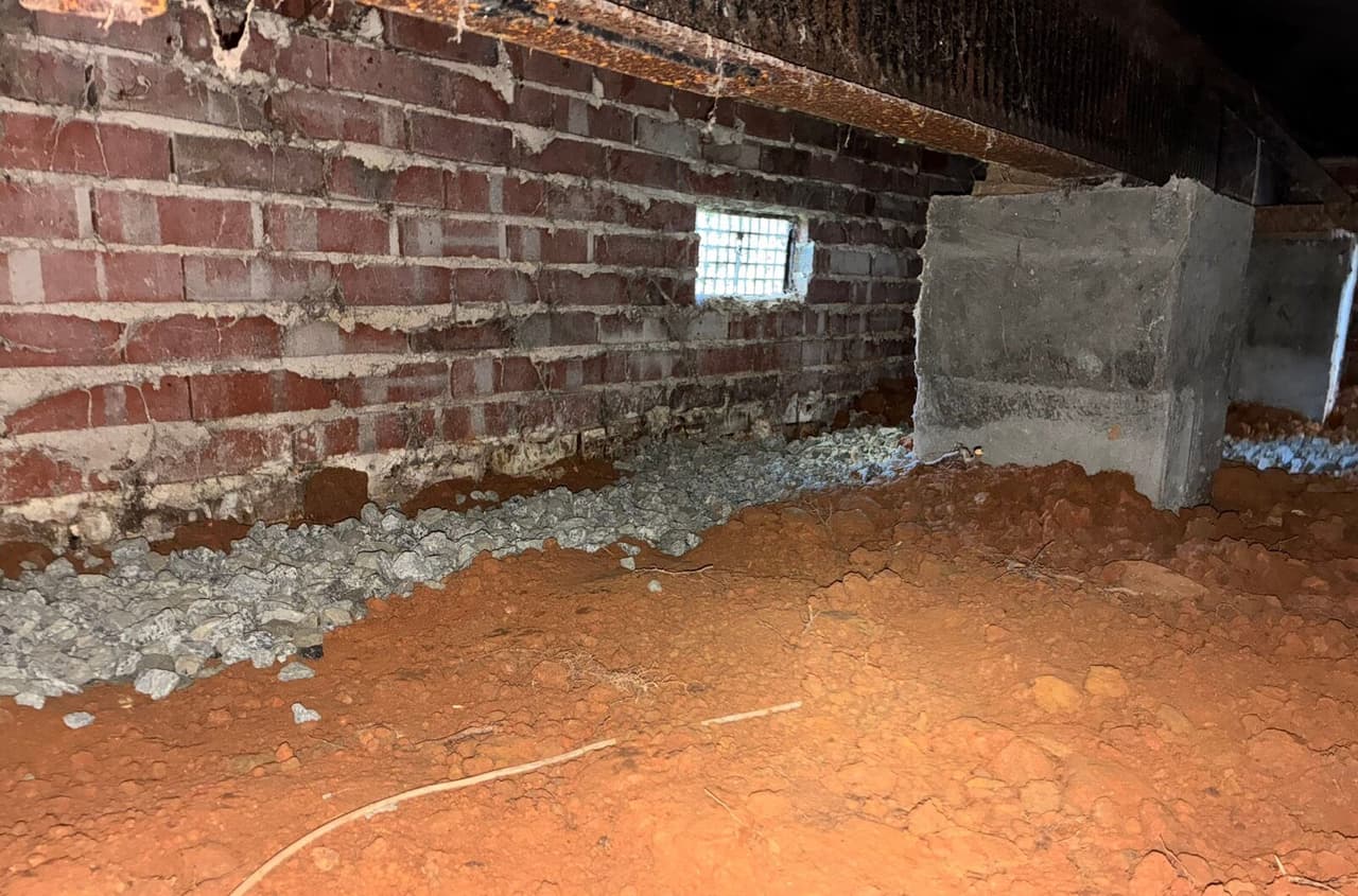 Gravel filled drainage trench running along a brick crawl space wall beside block support piers.