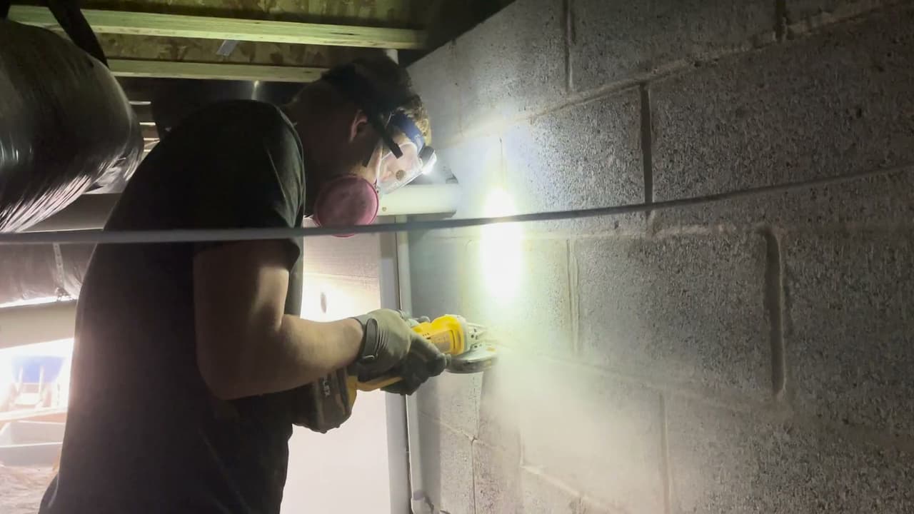 Technician grinding a cinder block wall for drainage system.