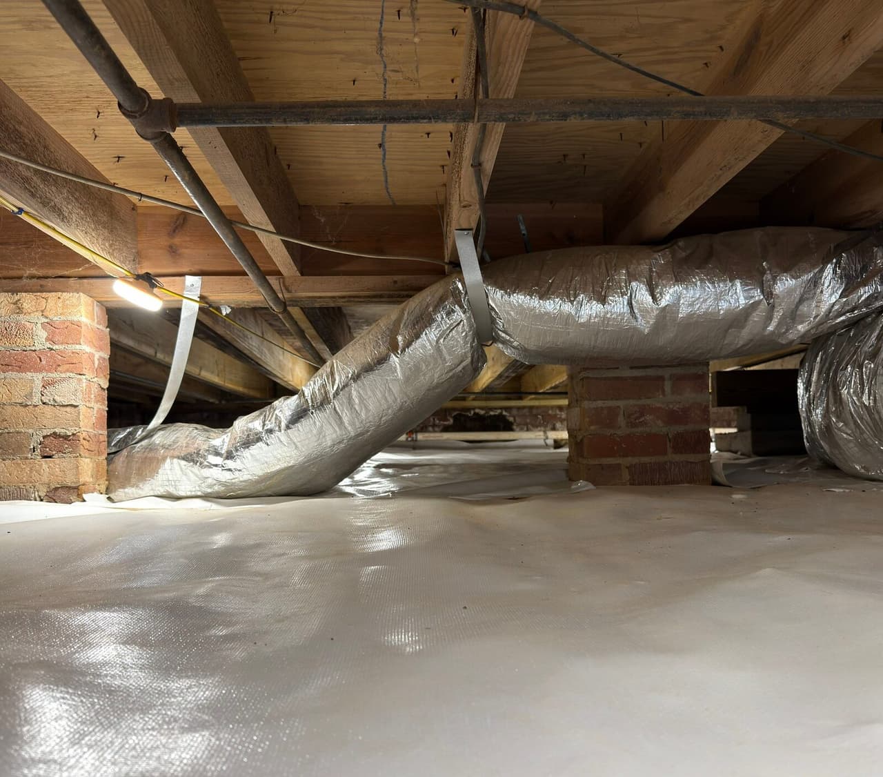 Crawl space with white liner across the floor, brick piers and silver insulated ducts hanging from joists.