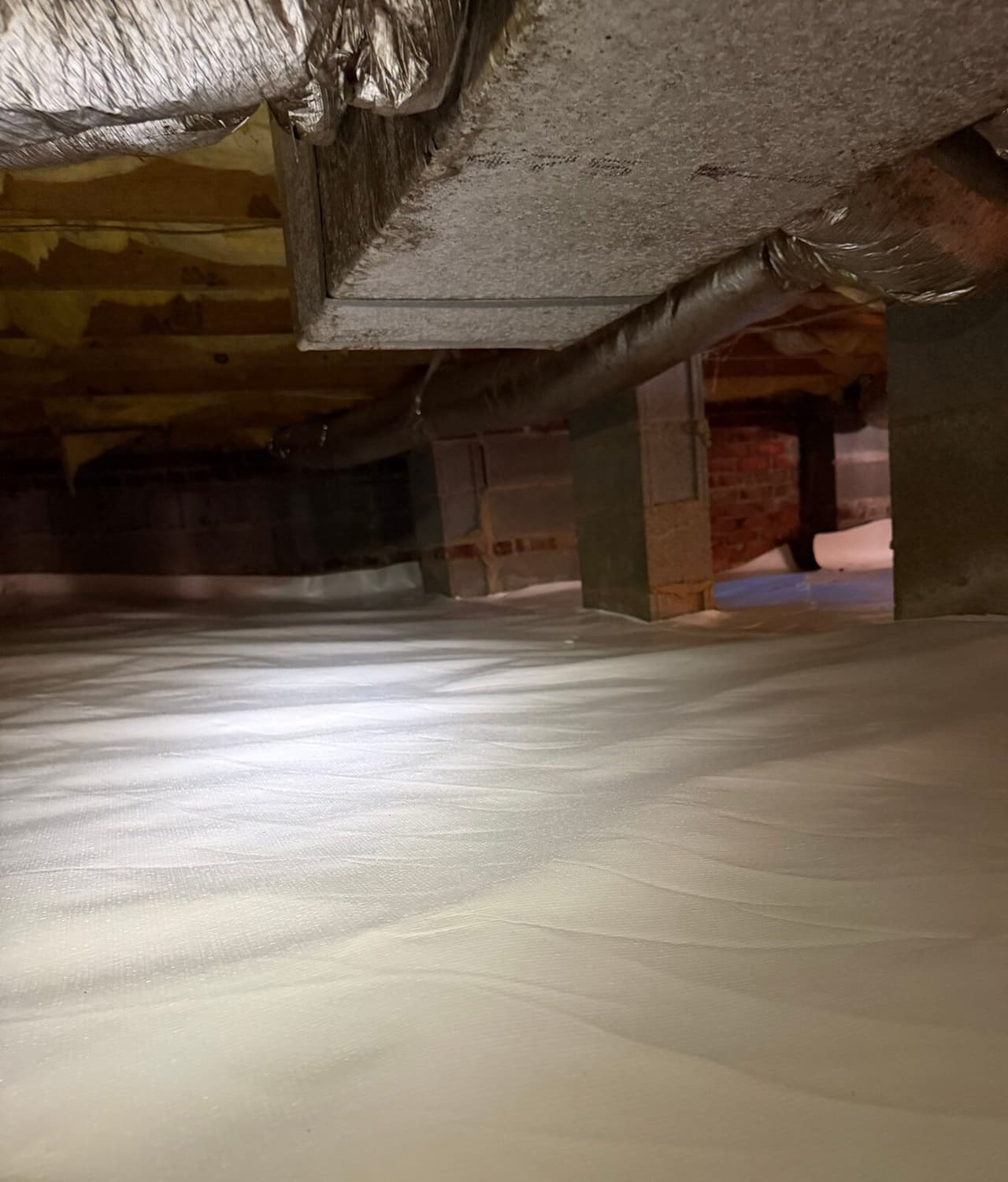 Low crawl space with white vapor barrier spread across the floor beneath a rectangular duct.
