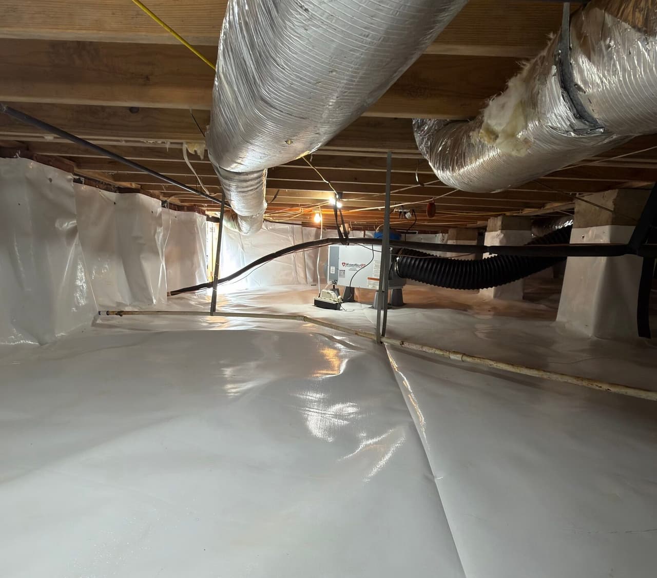 Lined crawl space with a dehumidifier set on the liner beneath ducts and joists.