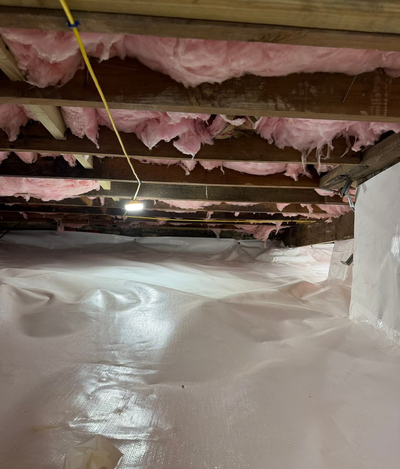 Long view of a crawl space floor covered in white liner under insulated joists and a hanging work light.
