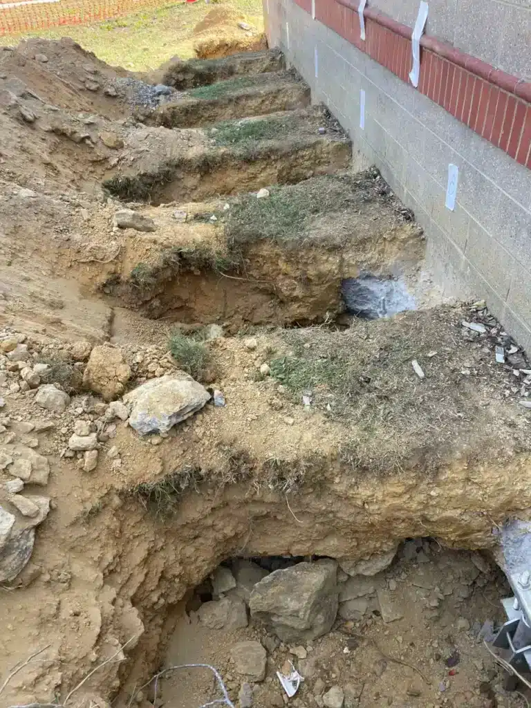 Stepped excavation beside a brick and block foundation wall with steel push piers installed for underpinning