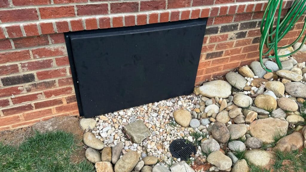 Black crawl space access door set in a brick foundation wall above river rock and a drain grate