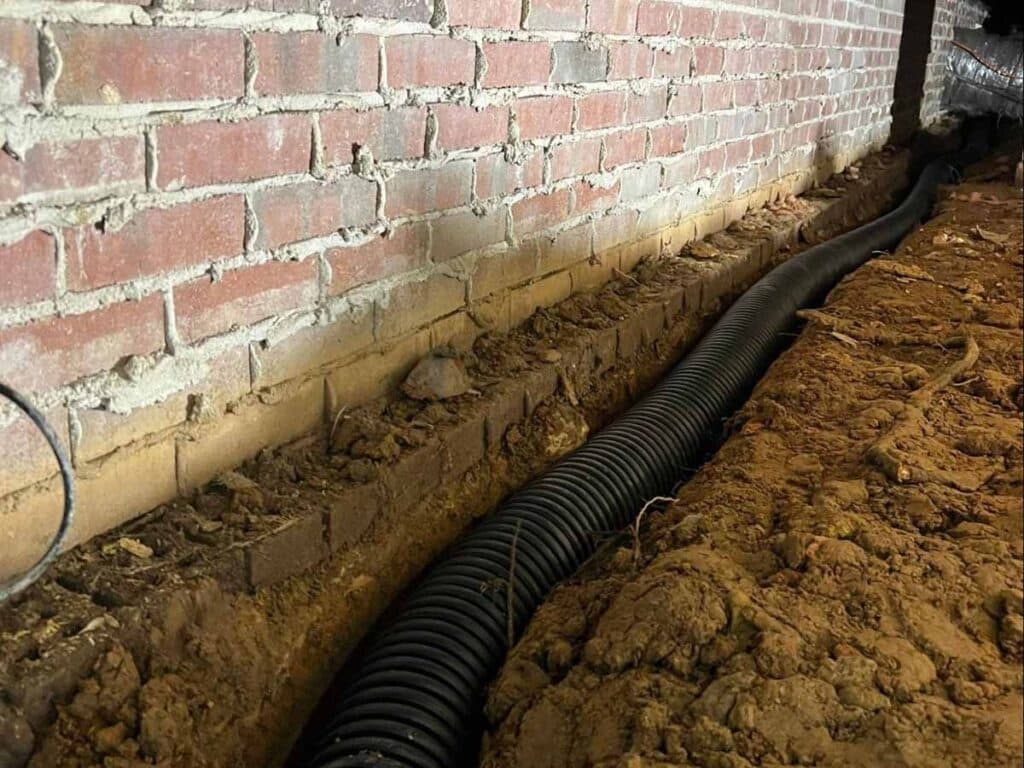 Black corrugated interior drain line laid in a trench along a brick crawl space wall